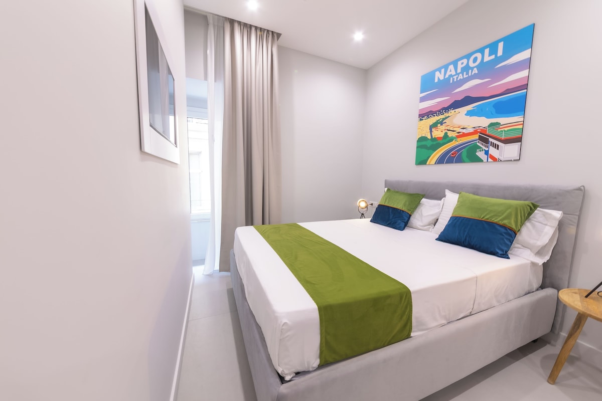 Een hotelkamer met een bed, nachtkastje, lamp, raam met gordijnen, en een schilderij van een stadsgezicht aan de muur.