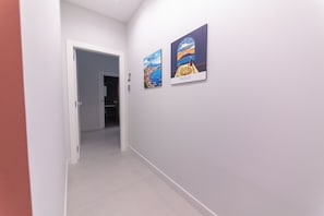 A hallway with a door leading to a room, two framed pictures on the wall, and a tiled floor. - The Ease (Naples)