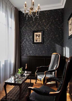 Une pièce aux couleurs sombres, agrémentée d’un lustre, d’un tableau encadré au mur, d’une table avec une bouteille et des verres, et de deux fauteuils rembourrés.