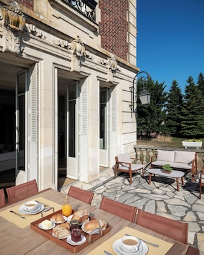 Un área de comedor al aire libre con suelo de piedra, mobiliario de madera y vista a un edificio de arquitectura clásica.