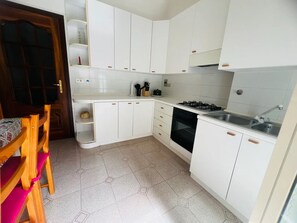 Cozinha com armários brancos, fogão a gás, pia e cadeira de madeira com almofada rosa.