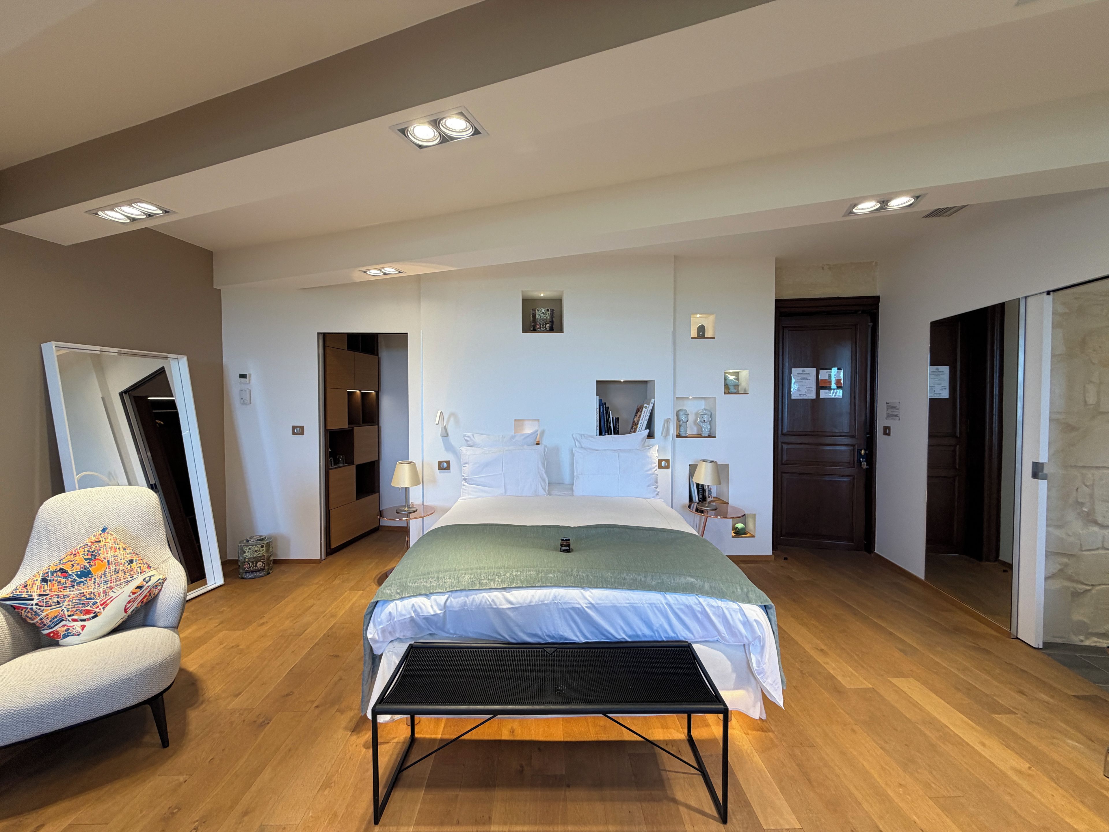 A modern hotel room with a large bed, a seating area with a chair, a bedside table, and a small table with a lamp.