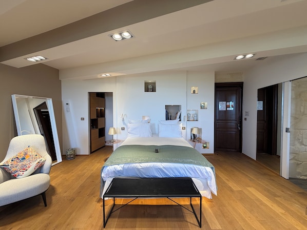 A modern hotel room with a large bed, a seating area with a chair, a bedside table, and a small table with a lamp.