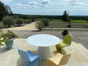 An outdoor seating area with a round table and colorful chairs, overlooking a landscaped garden and distant hills.