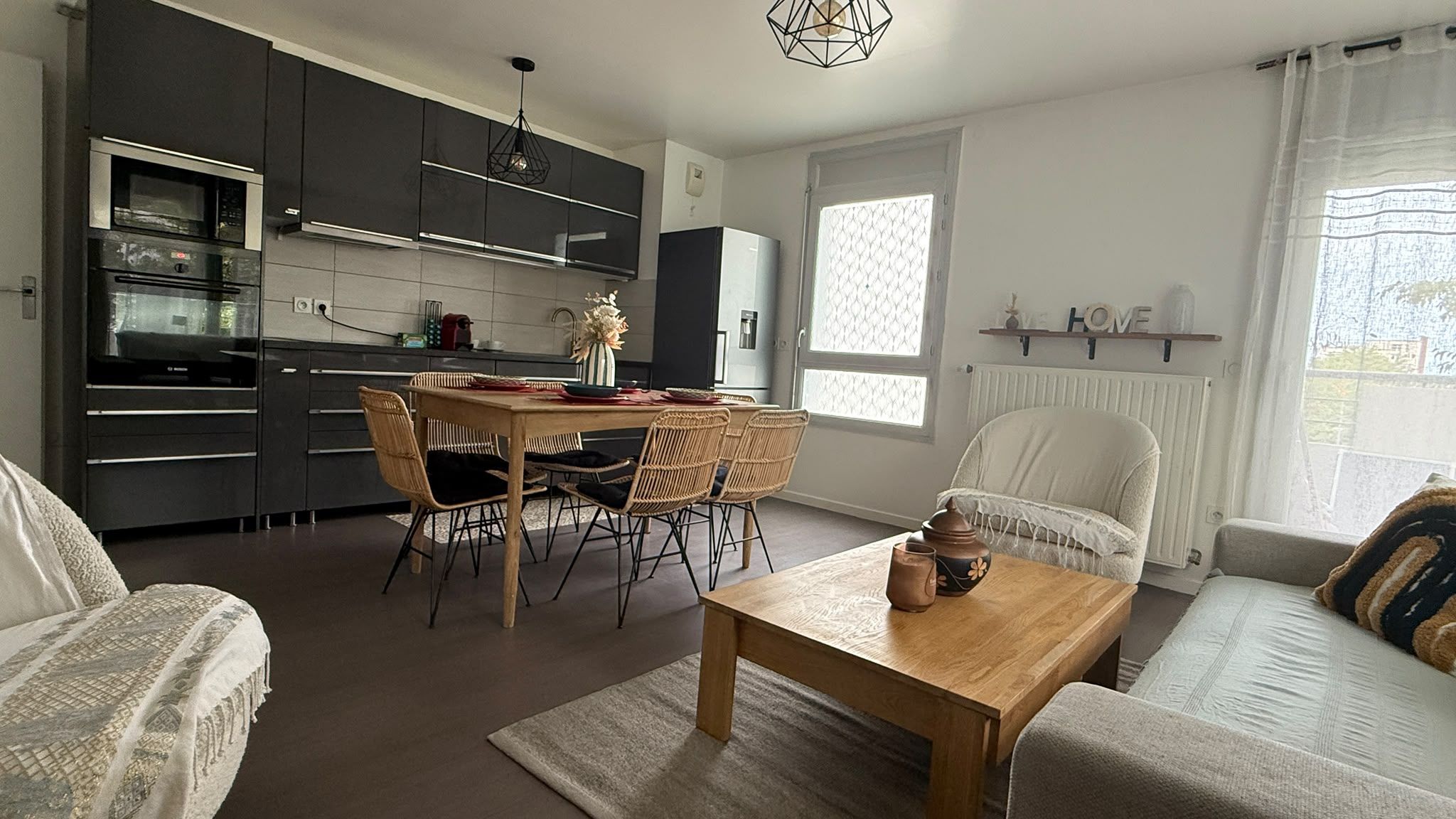 Une cuisine moderne avec des meubles de couleur sombre, un coin repas avec une table en bois et des chaises en osier, un canapé et une table basse.