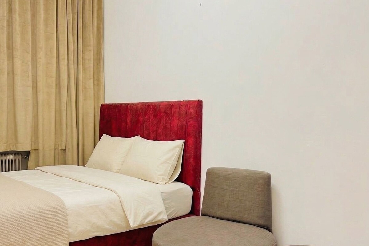 A bedroom with a bed, a red headboard, a round ottoman, and a small table.