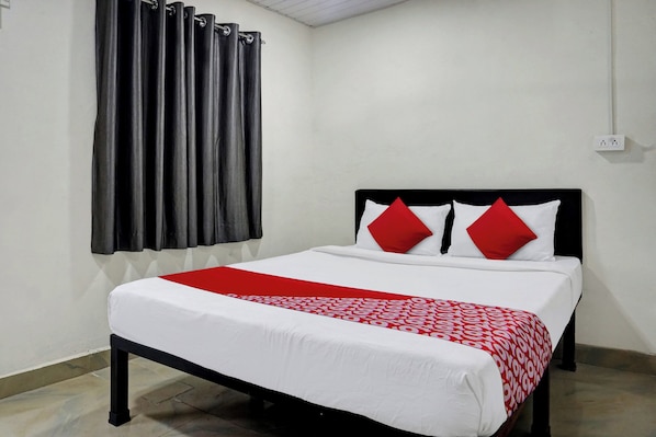 A single bed with a white and red bedspread, two red pillows, and a black headboard in a room with white walls and a window with grey curtains.