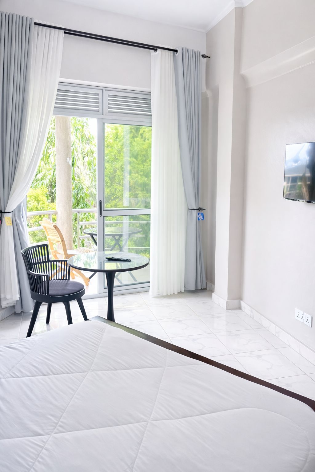 Une chambre d’hôtel avec un lit, une table ronde et une chaise, un balcon avec des rideaux et un téléviseur fixé au mur.