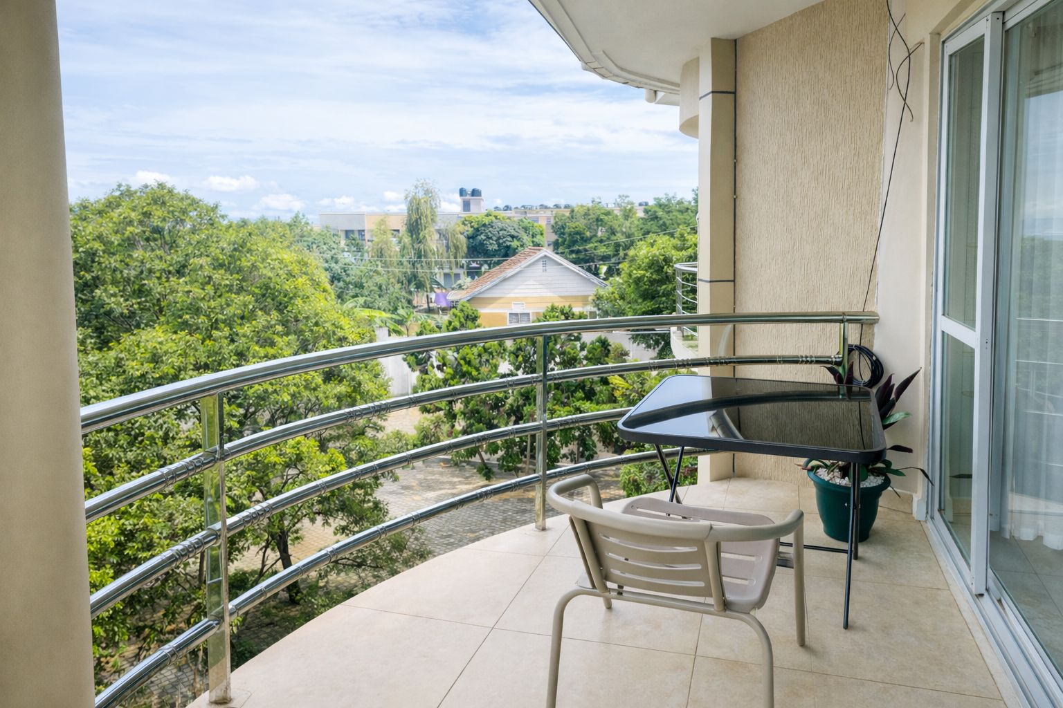 A balcony with a table and chair, a potted plant, and a view of greenery and buildings.