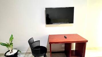 Quarto com cama, escrivaninha, cadeira, televisão fixada na parede e planta em vaso.