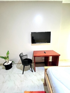 Quarto com cama, escrivaninha, cadeira, televisão fixada na parede e planta em vaso.