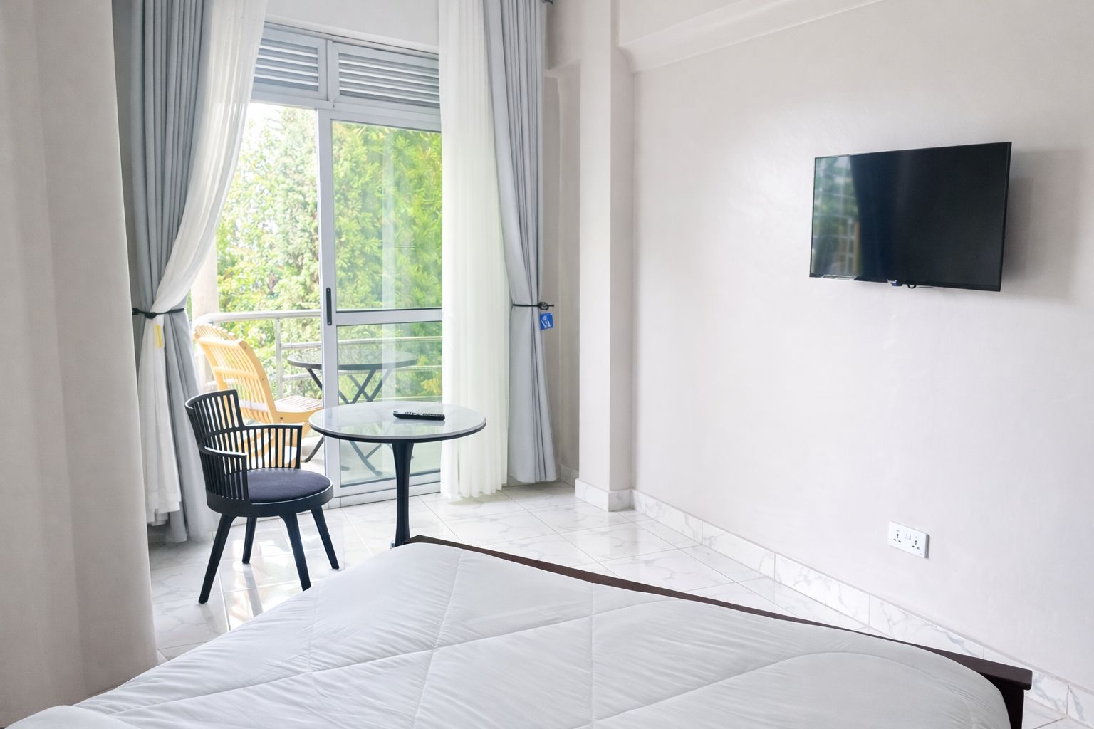 Quarto de hotel com cama, televisão na parede, varanda com mesa e cadeiras, e vista para o verde lá fora.