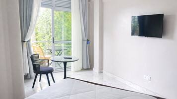 Quarto de hotel com cama, televisão na parede, varanda com mesa e cadeiras, e vista para o verde lá fora.