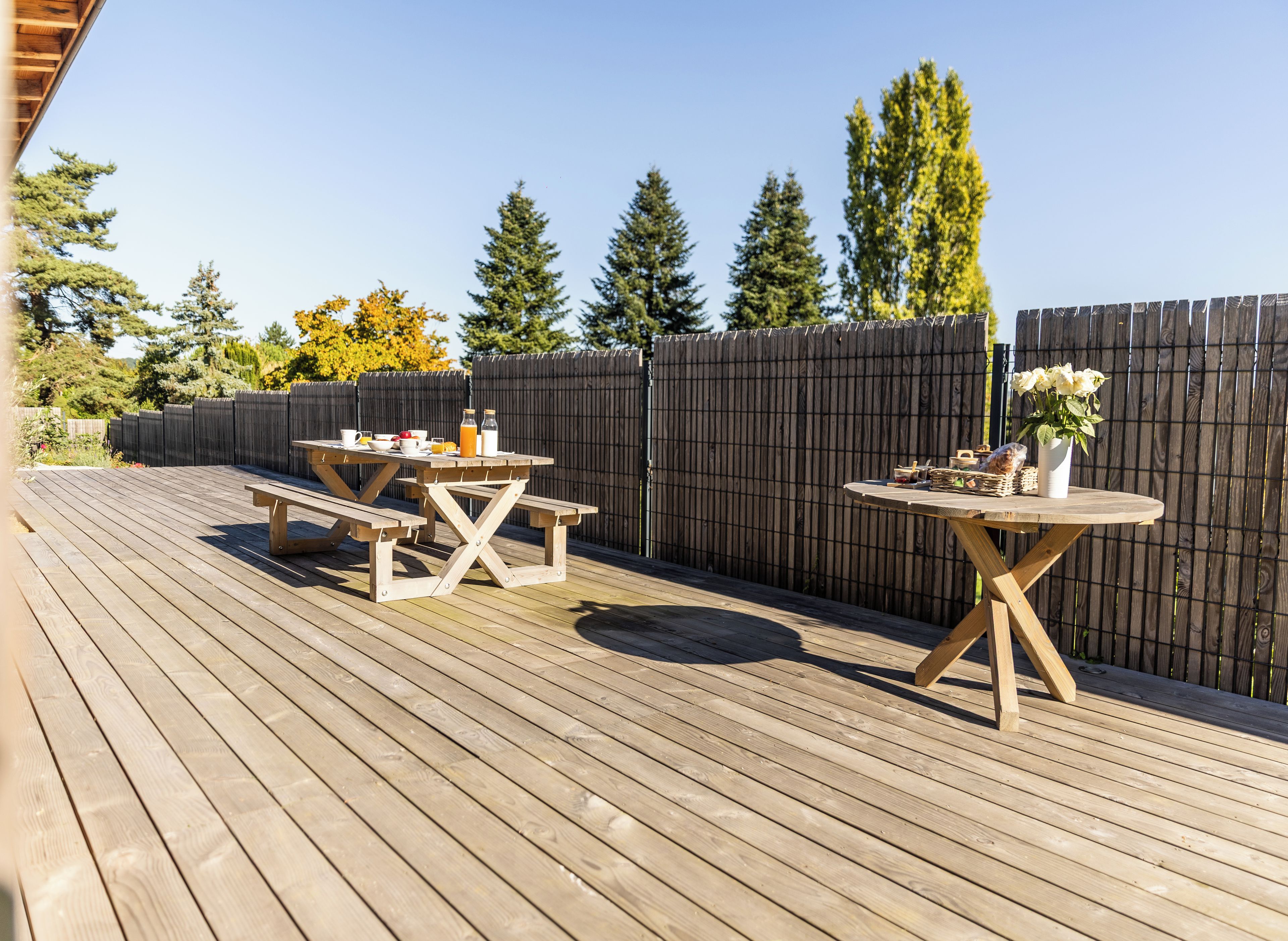 Una terraza de madera con mesas de picnic y una mesa redonda, rodeada por una valla de madera y árboles.