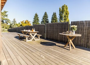 Una terraza de madera con mesas de picnic y una mesa redonda, rodeada por una valla de madera y árboles.
