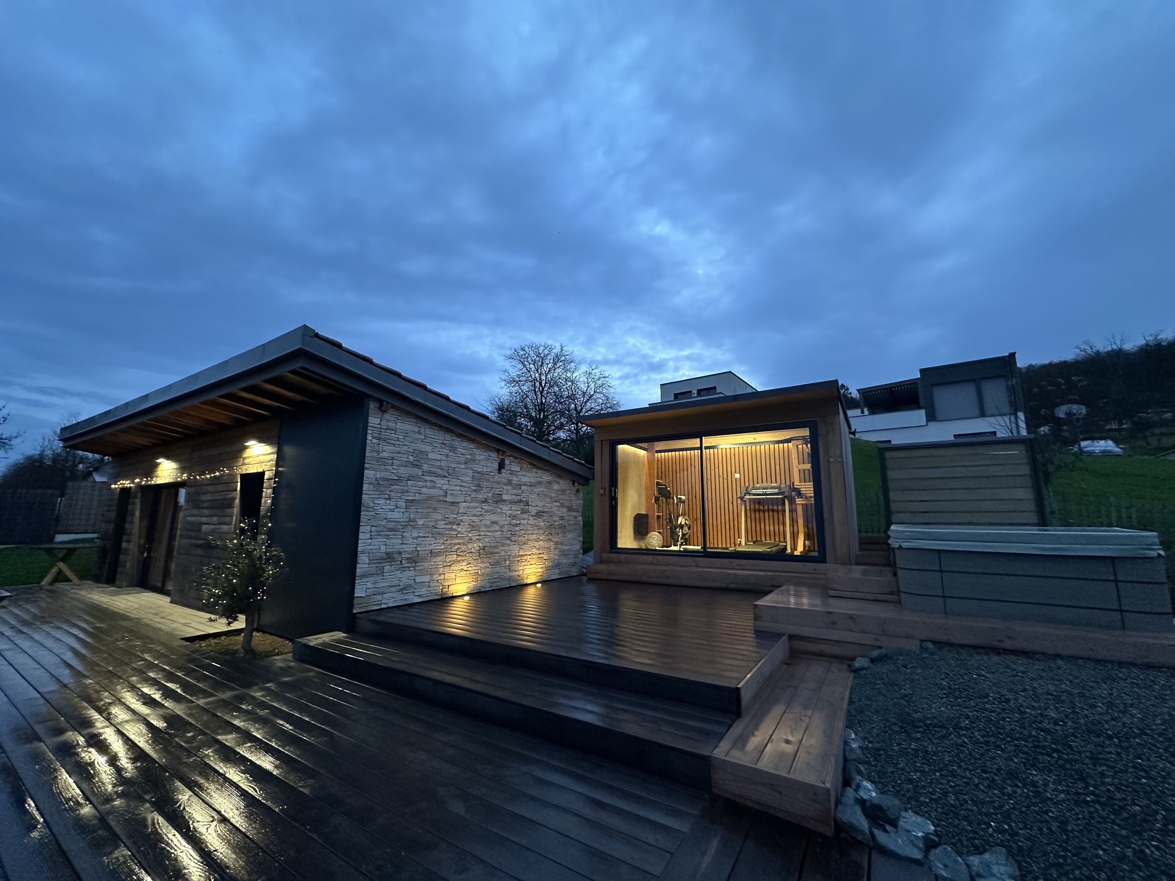 Una casa moderna con fachada de piedra, terraza de madera y zona de porche con paredes de vidrio.