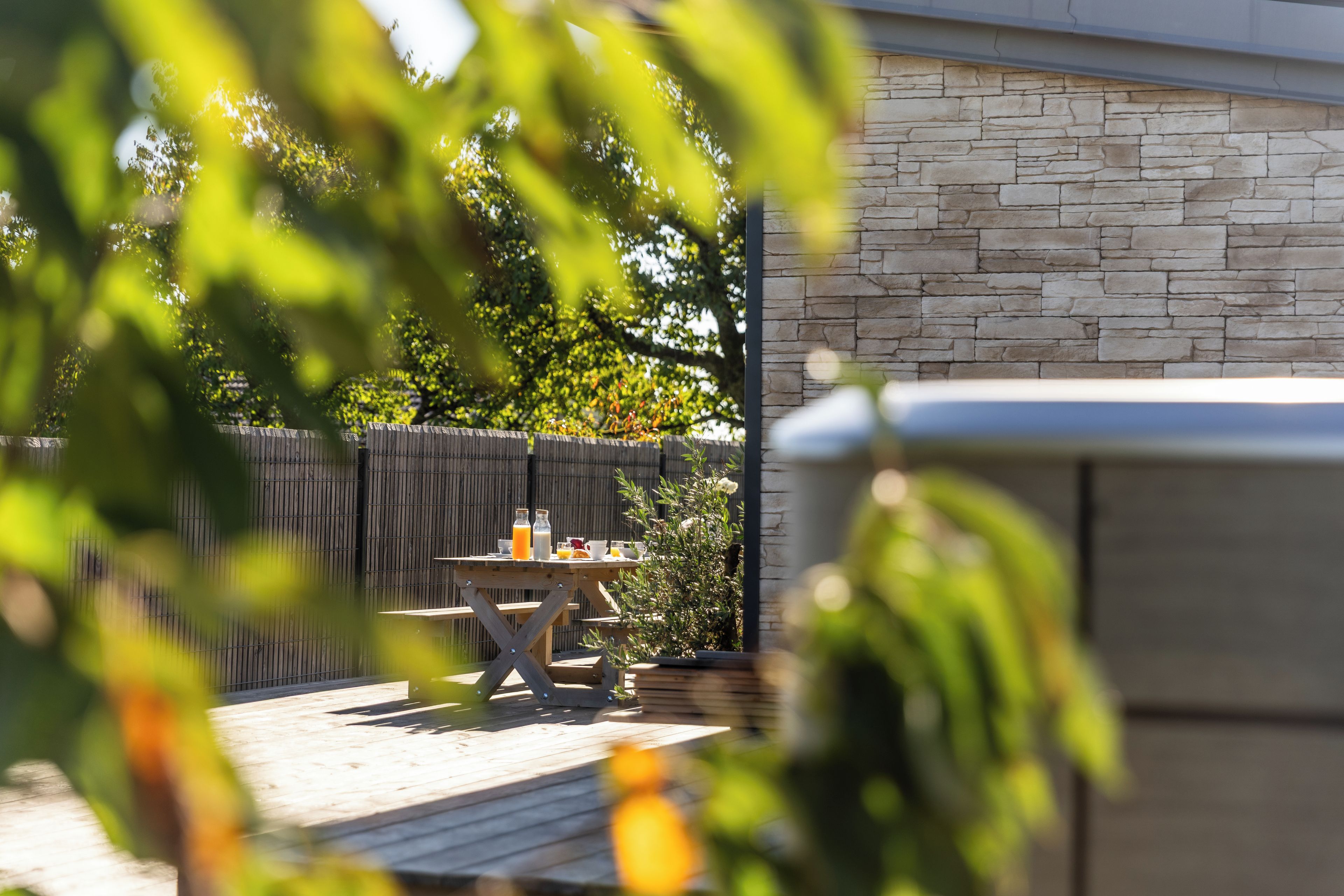 Una terraza de madera con una mesa dispuesta para el desayuno, un muro de piedra y una valla de madera al fondo.