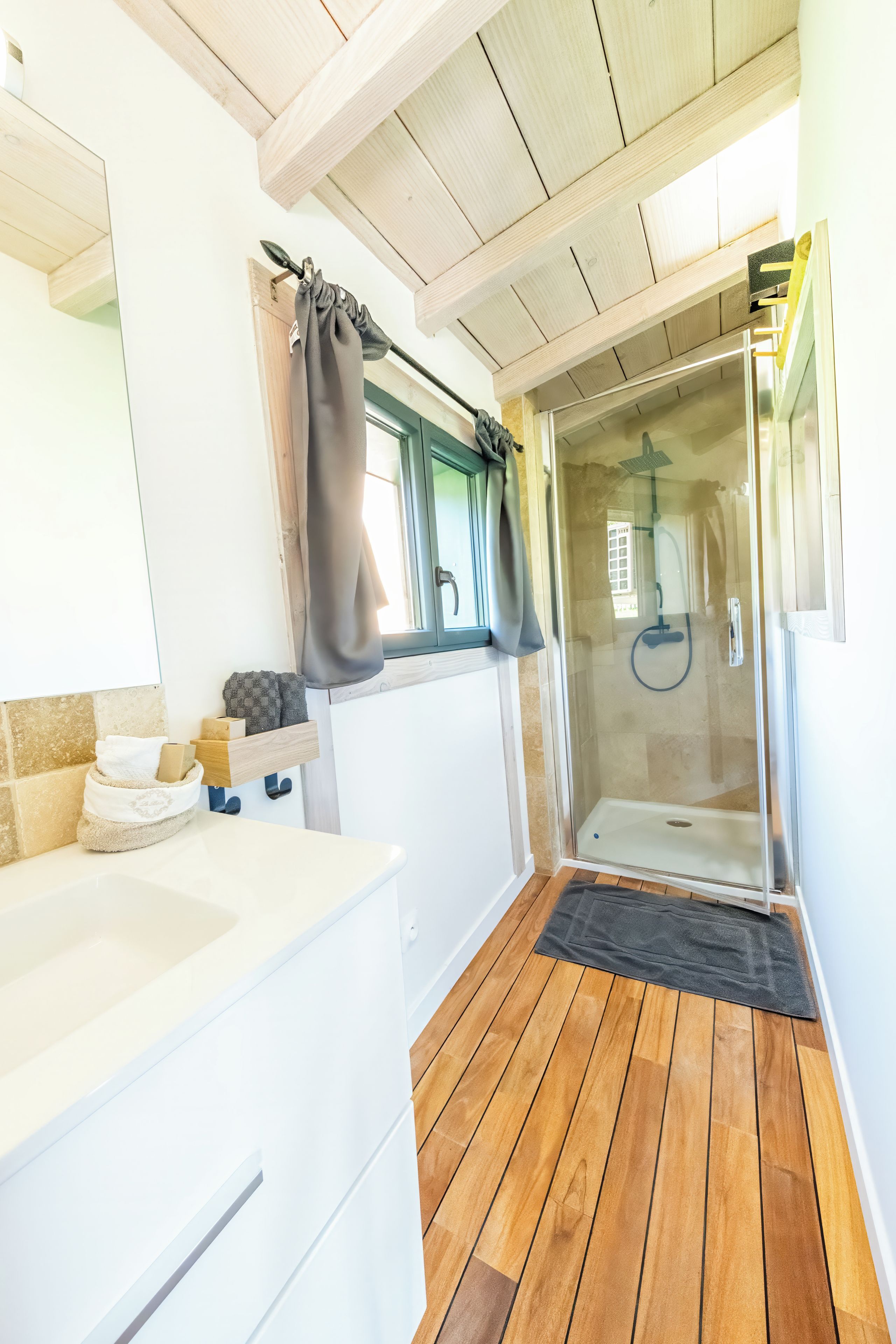 Un baño moderno con piso de madera, un lavamanos blanco, cabina de ducha con paredes de vidrio y una ventana con cortinas.