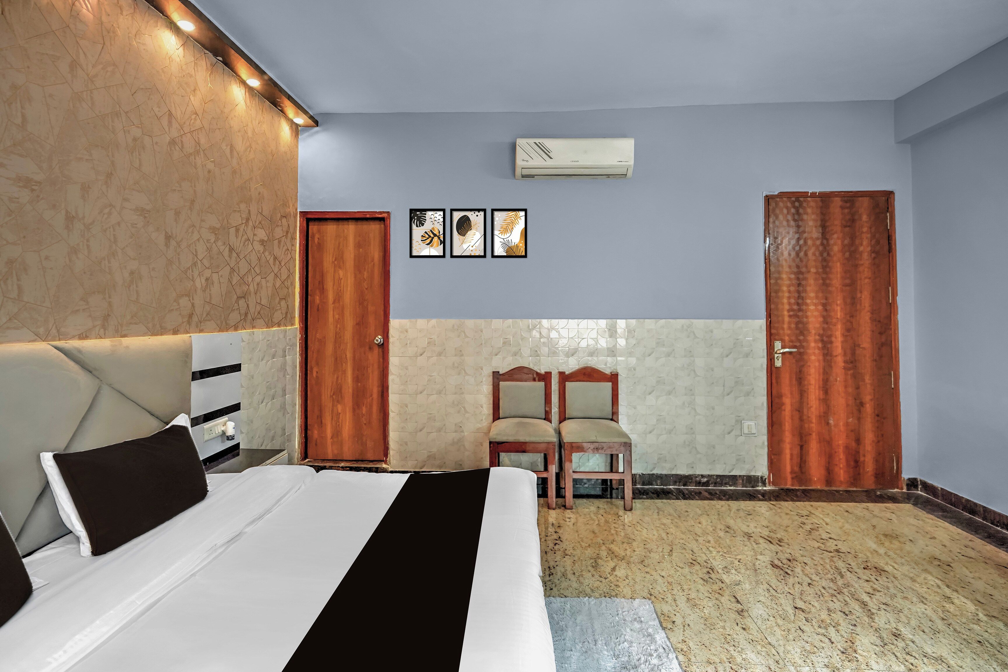 A hotel room with a bed, two wooden doors, a wall-mounted air conditioner, and three framed pictures.