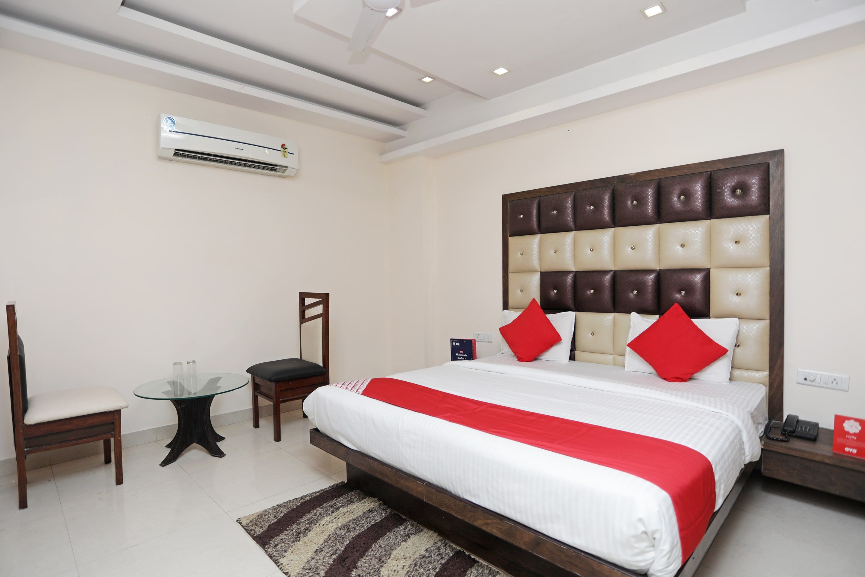 A hotel room with a large bed, a chair, a small table, and an air conditioning unit.