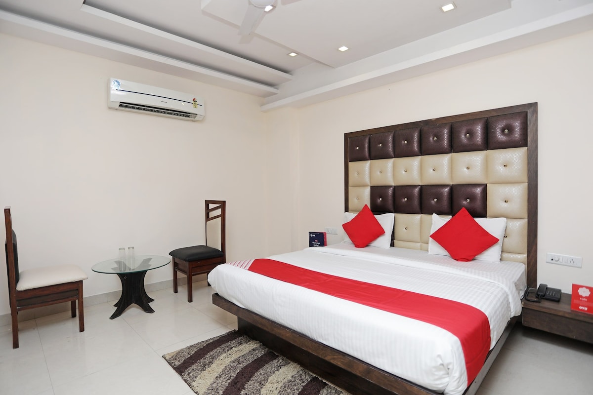 A hotel room with a large bed, a chair, a small table, and an air conditioning unit.
