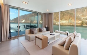 Una sala de estar moderna con un amplio ventanal corredera que da a un balcón con vistas a un cuerpo de agua y colinas.