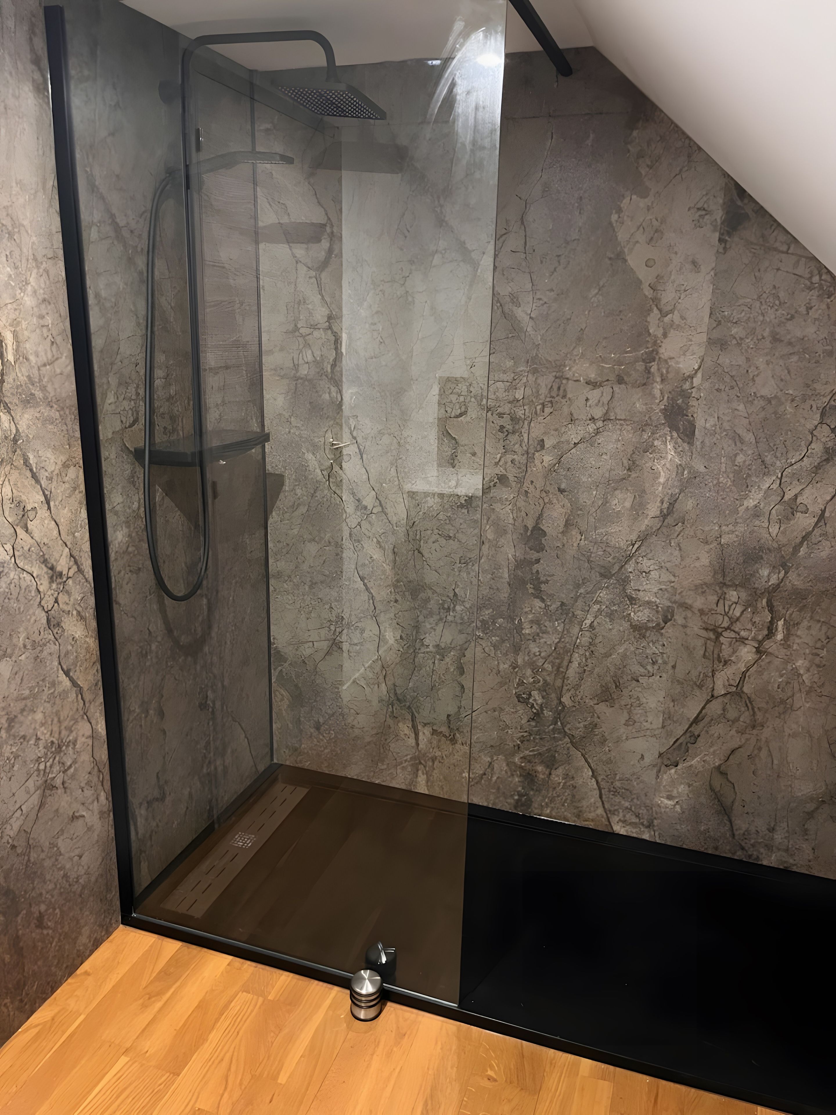 Eine gläserne Dusche mit Handbrause, Holzboden und Marmorwand.