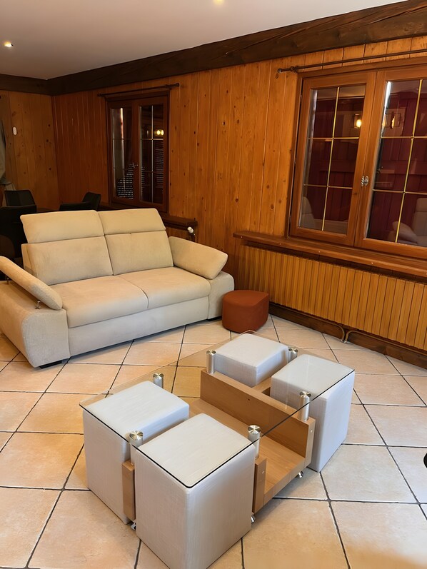 Een moderne woonkamer met een beige bank, een houten salontafel en een groot raam met houten jaloezieën.
