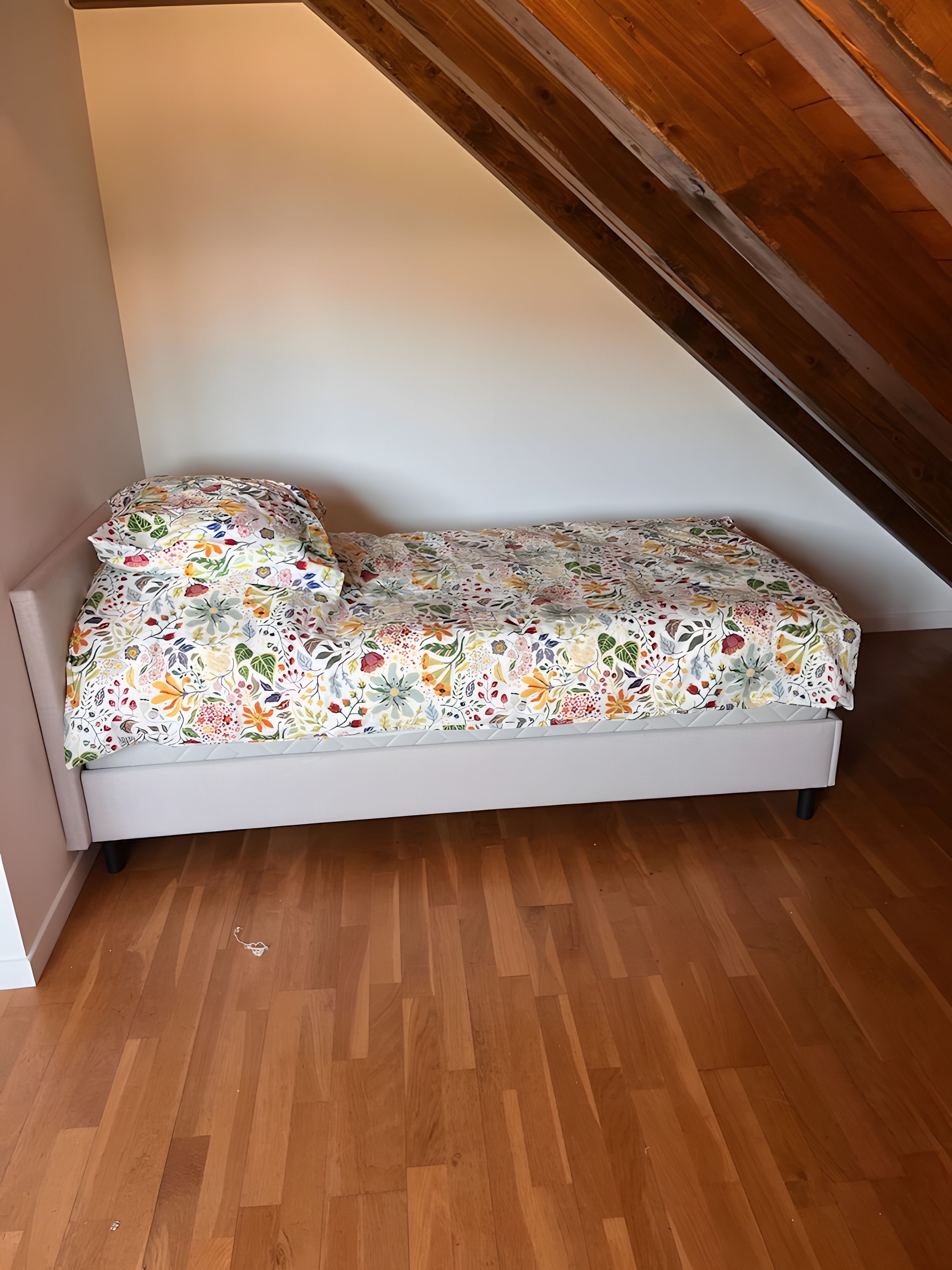 Una cama pequeña con colcha floral en una habitación con piso de madera y techo inclinado.