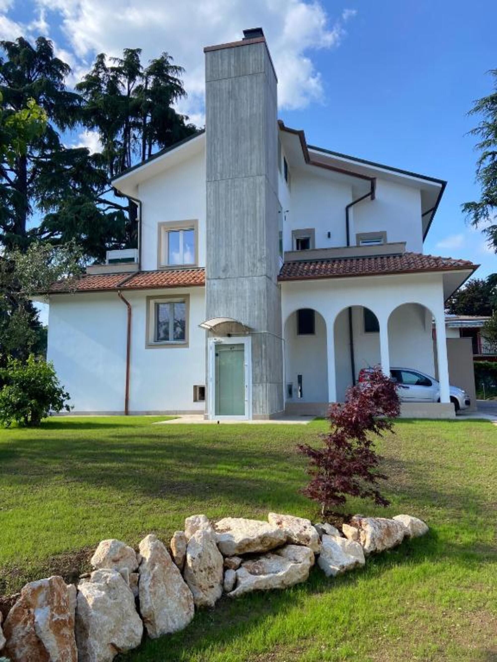 Una casa a due piani con un camino centrale, un portico coperto e un muro di pietra nel giardino.