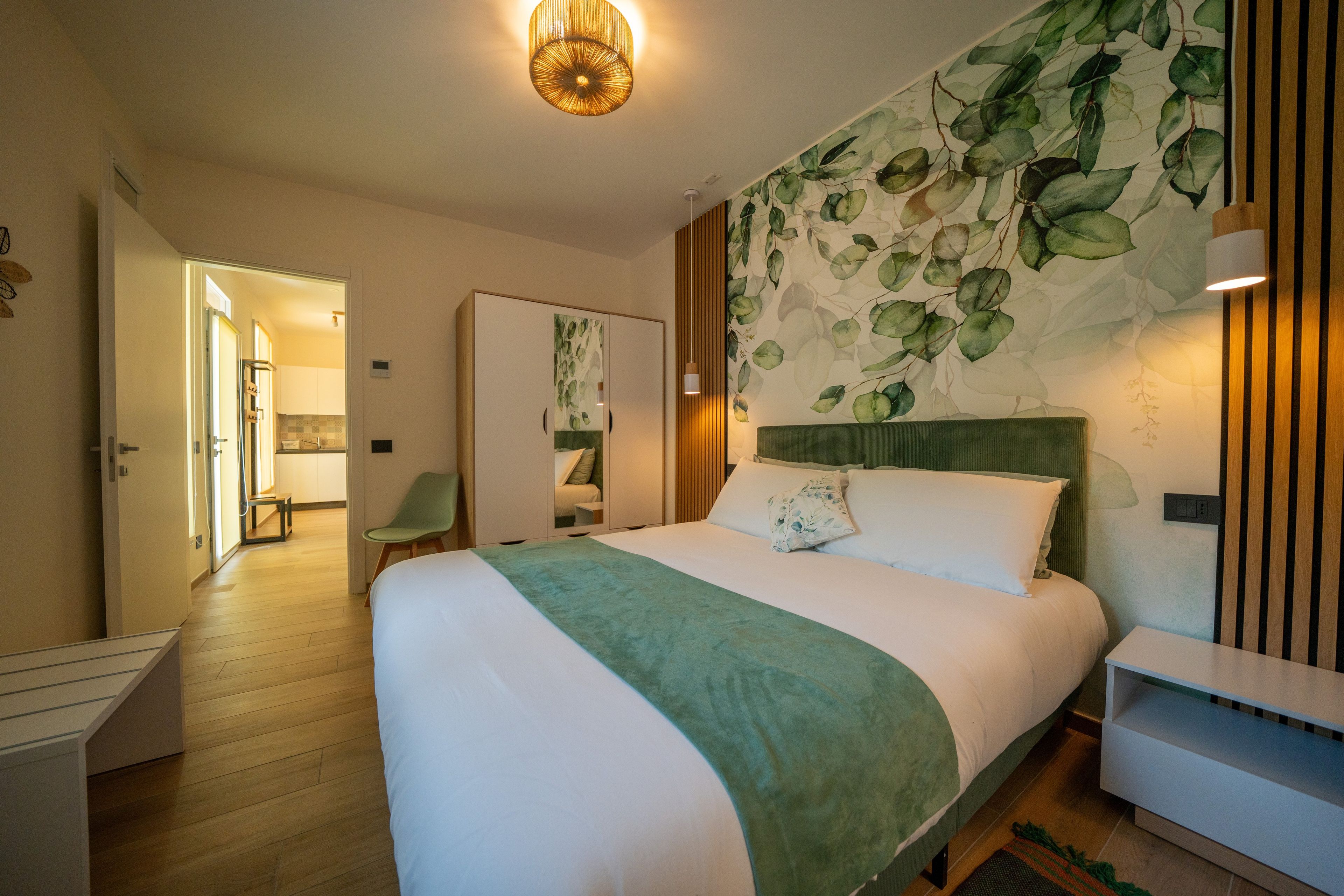Camera d'hotel con un letto grande, un poggiatesta verde, una parete decorata con un motivo a foglia, un comodino in legno e una porta che conduce al bagno.