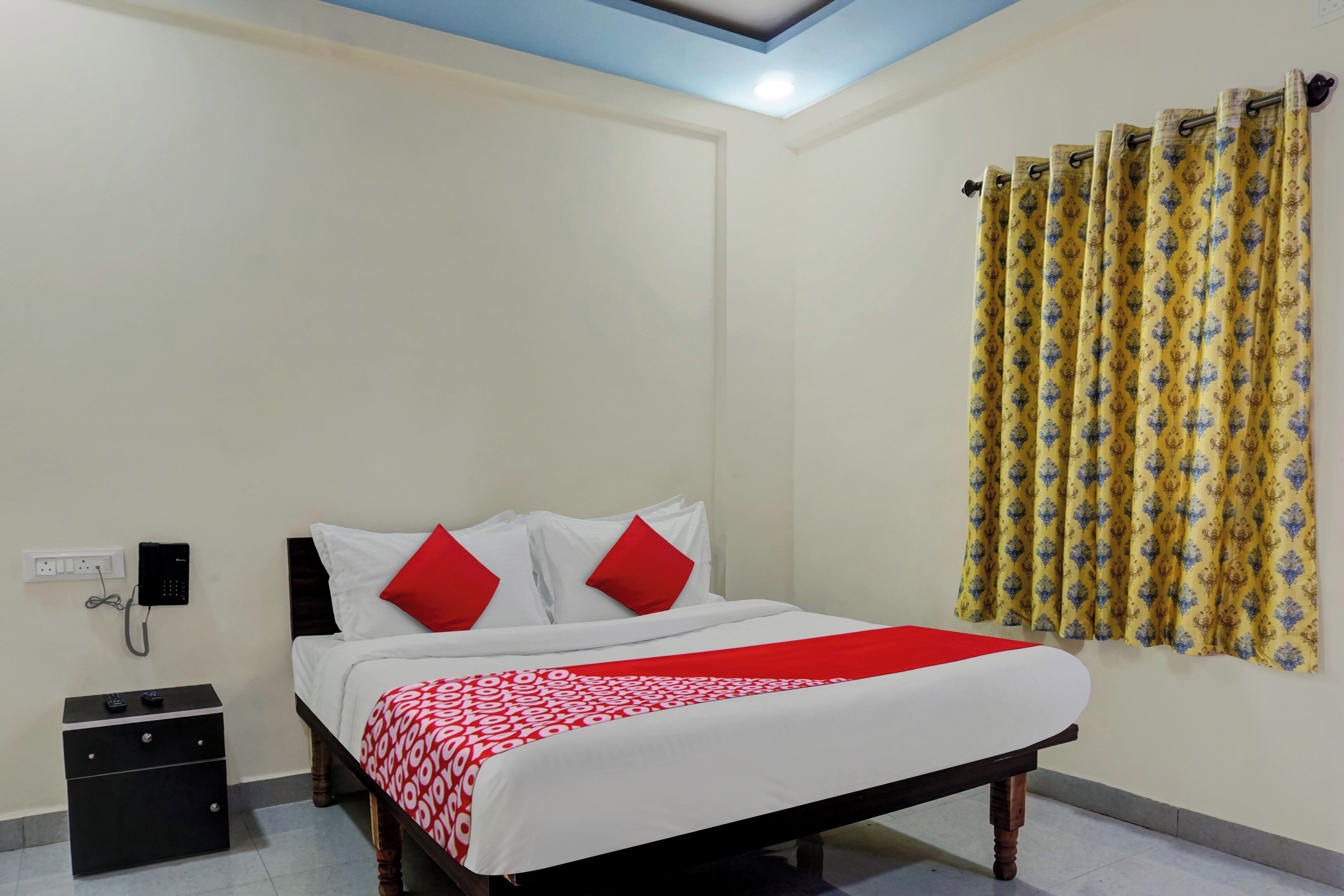 A hotel room with a bed, a bedside telephone, a small cabinet, and a curtain with a pattern.