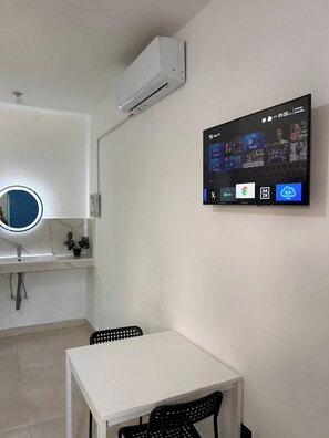 Habitació amb taula blanca, dues cadires, aire condicionat fixat a la paret, televisió que mostra contingut esportiu i un mirall rodó amb llum.