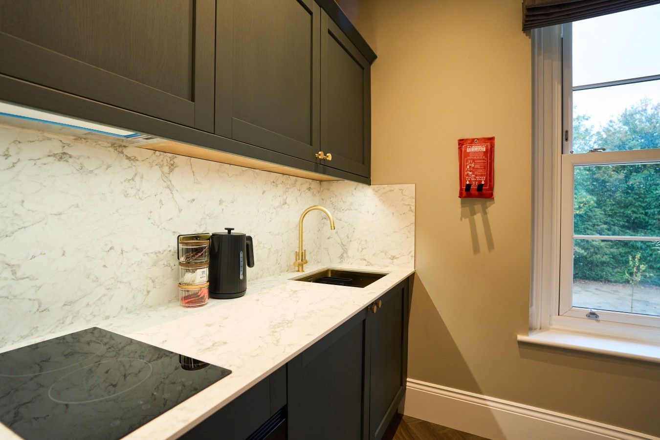 A kitchen with dark cabinets, a marble countertop, a black coffee maker, and a window with a view of greenery.