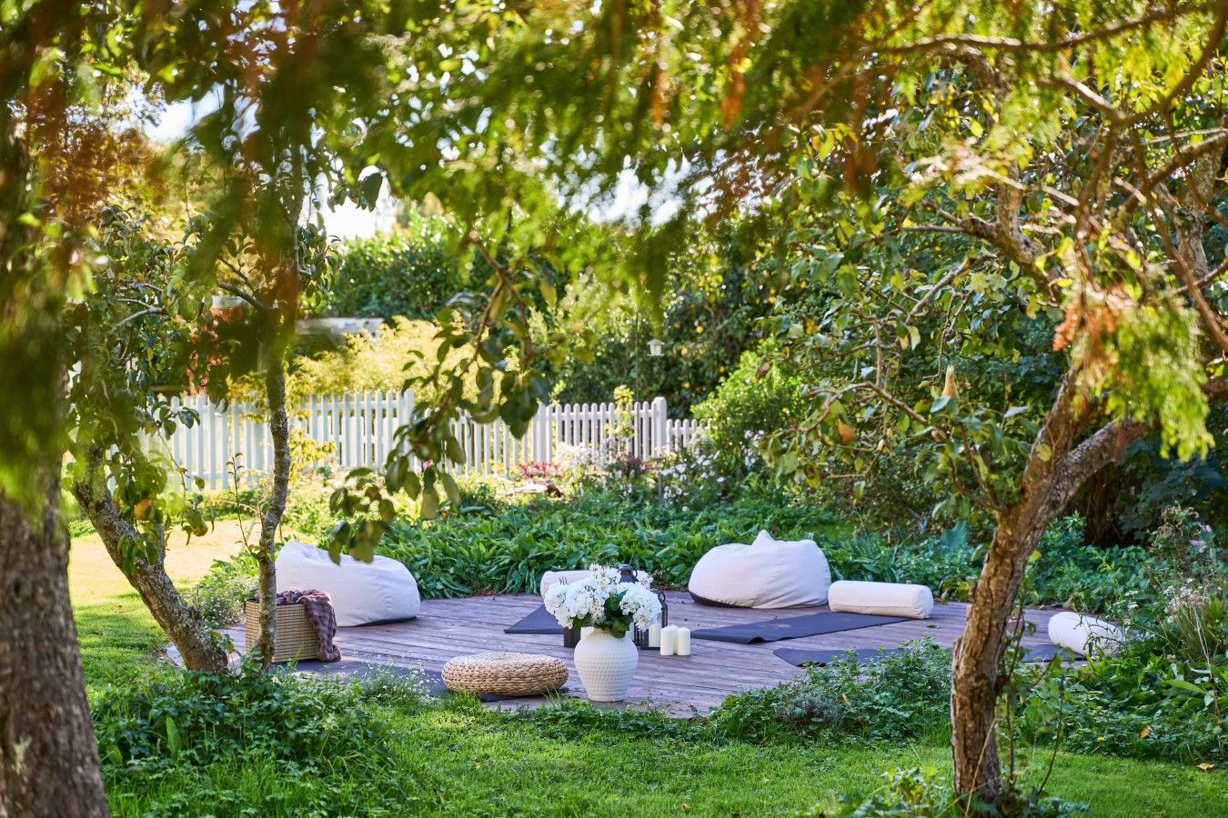 A garden with a wooden deck, white cushions, a woven basket, and a white vase with flowers.