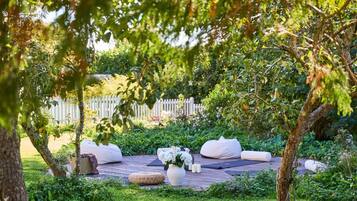 A garden with a wooden deck, white cushions, a woven basket, and a white vase with flowers.