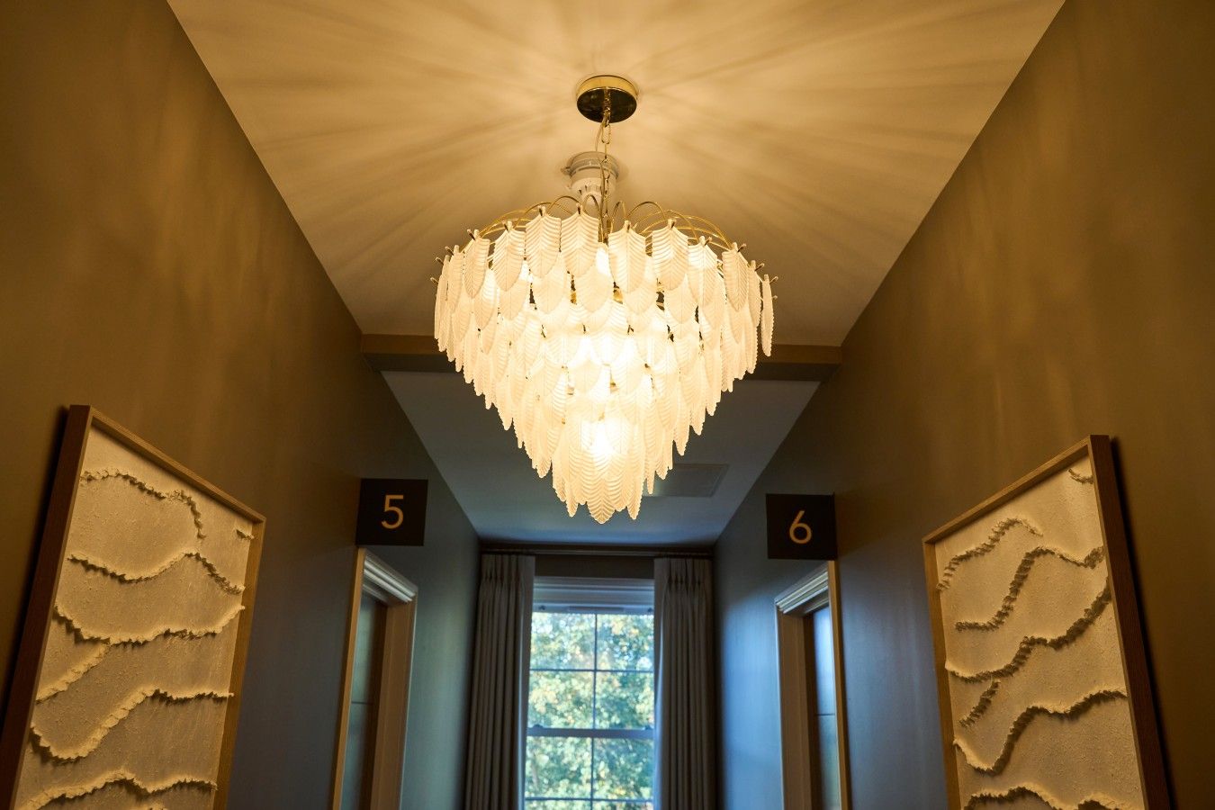 A hallway with a chandelier, numbered doors, and a window with a view of trees.
