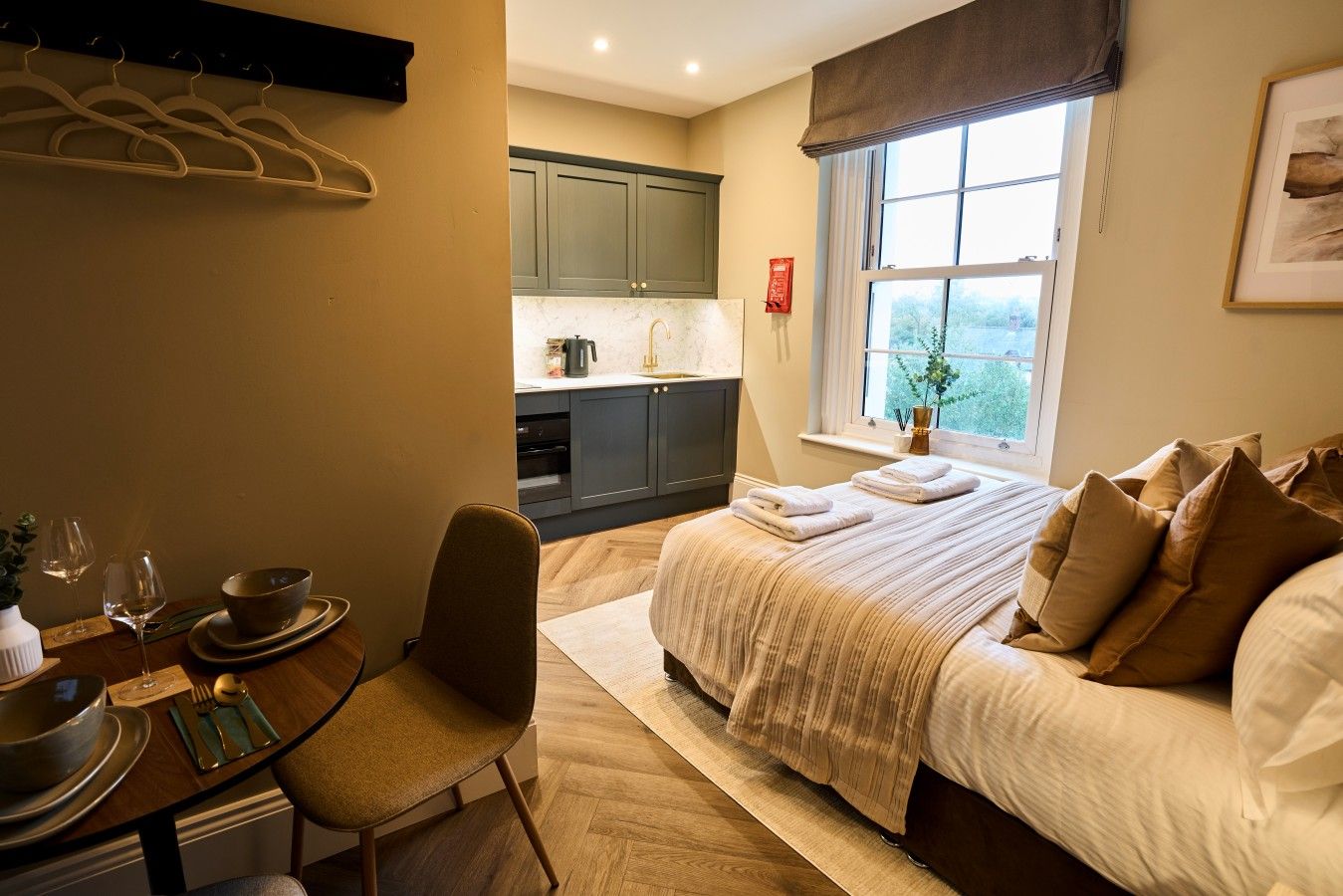 A hotel room with a bed, a dining table with chairs, a kitchen area, and a window with a view.