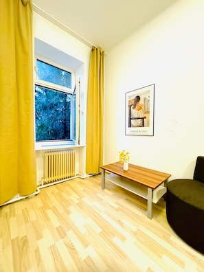 Une pièce avec une fenêtre, un radiateur, un plancher en bois, une petite table sur laquelle se trouve un vase, et un tableau encadré accroché au mur.