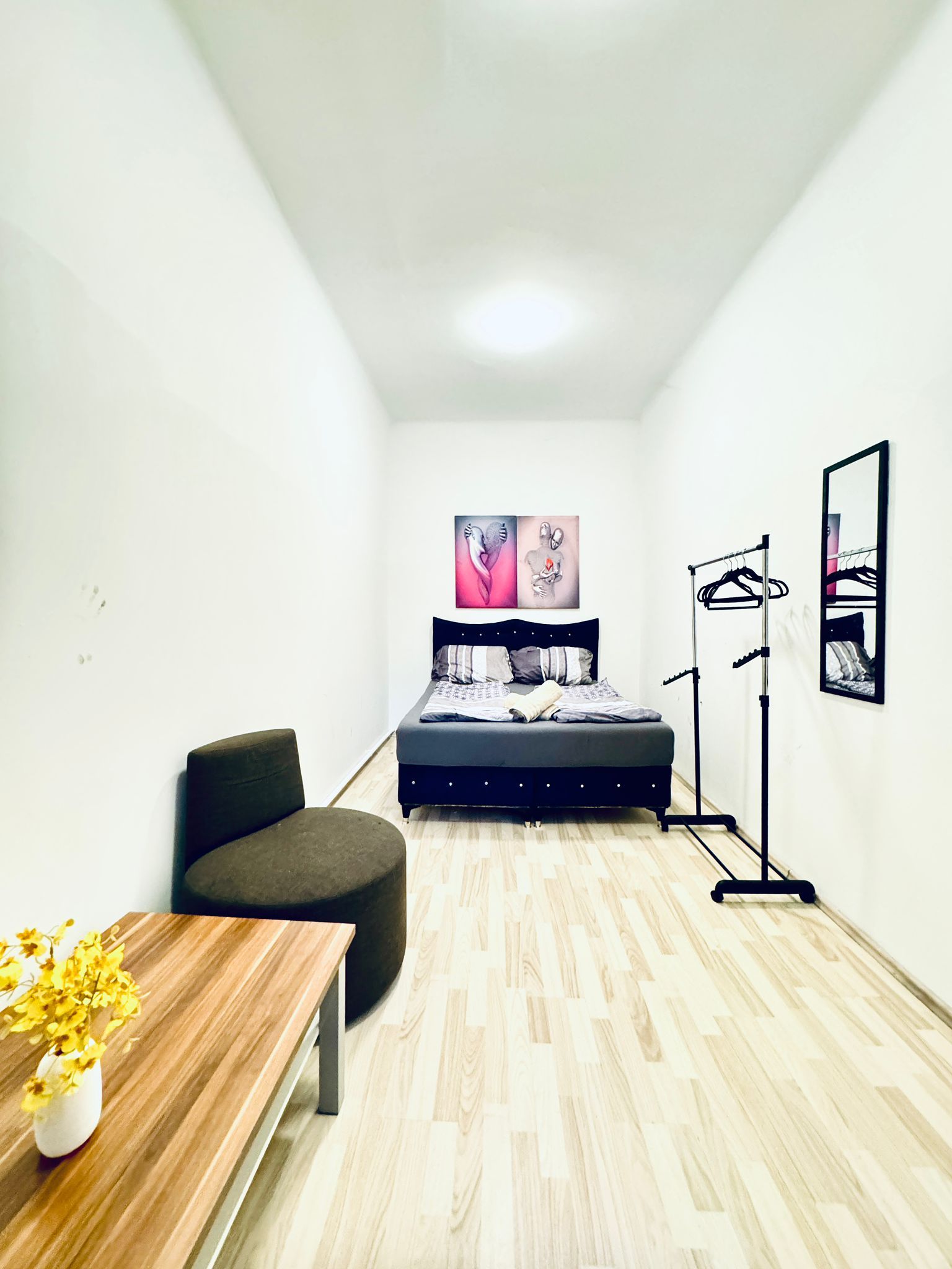 A small, modern room with a bed, a chair, a table with flowers, and a mirror on the wall.