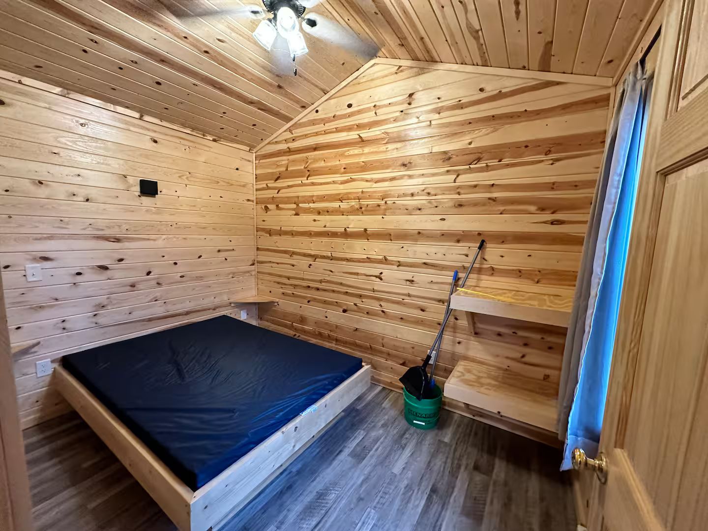 A small, wooden cabin room with a single bed, a ceiling fan, and a door leading outside.