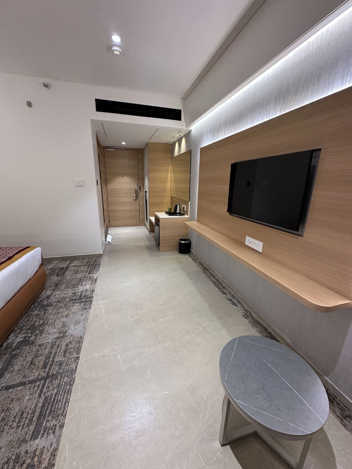 A modern hotel room with a bed, a bench, a TV mounted on the wall, and a small desk with a mirror.