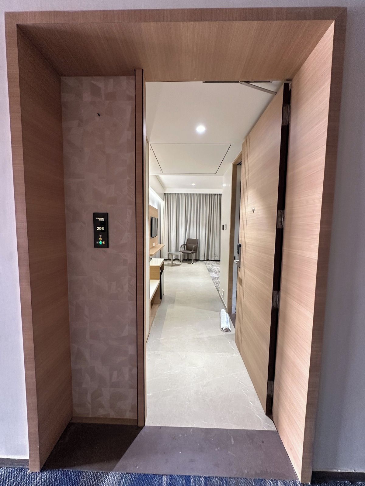 A hotel corridor with a wooden door, a digital elevator control panel, a seating area with a table and chairs, and a view into a room with a bed and curtains.