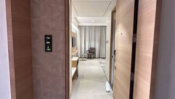 A hotel corridor with a wooden door, a digital elevator control panel, a seating area with a table and chairs, and a view into a room with a bed and curtains.