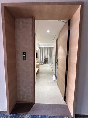 A hotel corridor with a wooden door, a digital elevator control panel, a seating area with a table and chairs, and a view into a room with a bed and curtains. - Imperia by The Sky Imperial Somnath (Keshod)