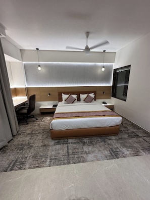 A modern hotel room with a large bed, a desk, a chair, a ceiling fan, and a window. - Imperia by The Sky Imperial Somnath (Keshod)