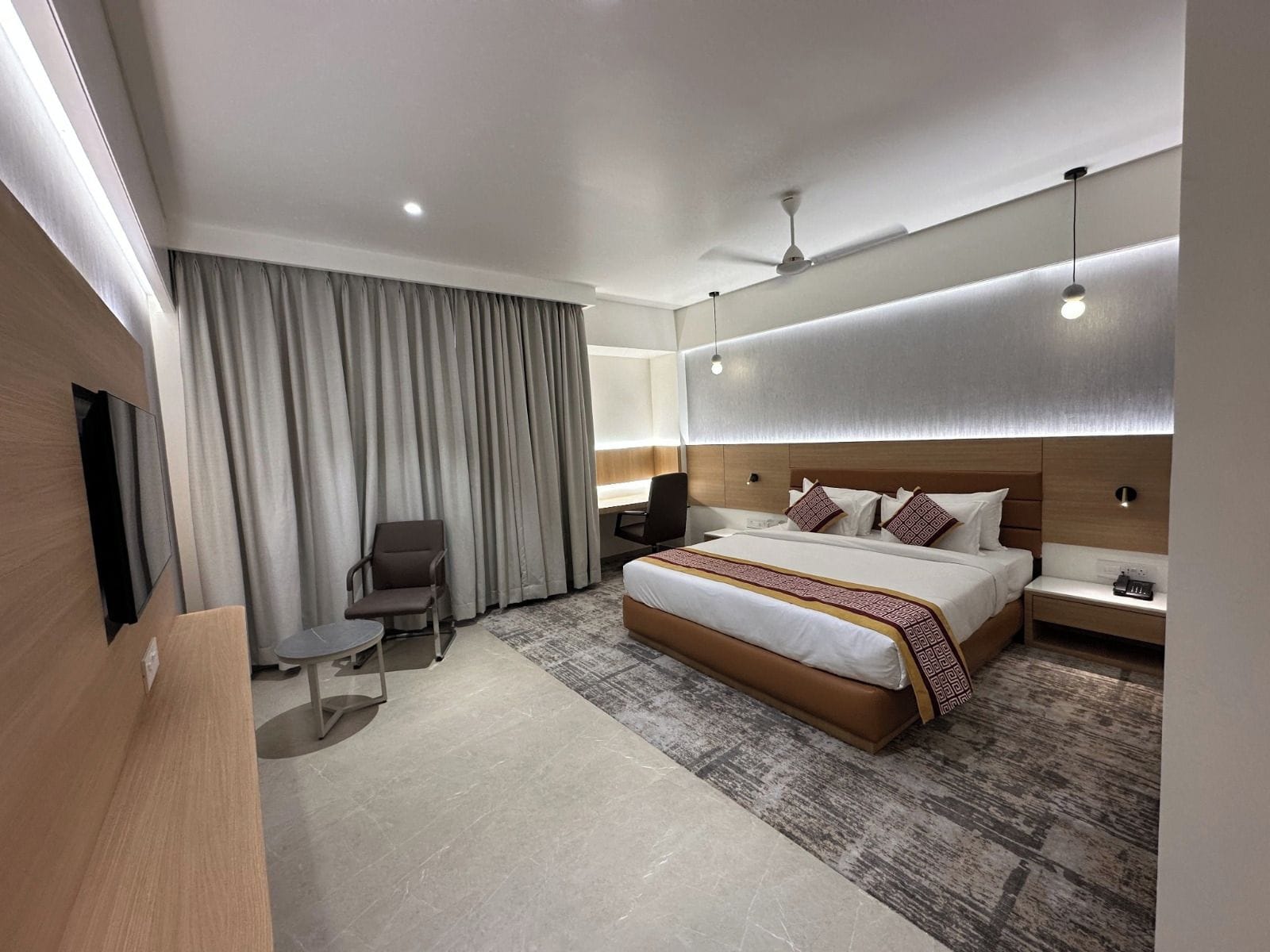 A hotel room with a large bed, a desk, a chair, a TV, and a ceiling fan.