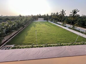 A large, well-maintained grassy area with a paved walkway, surrounded by palm trees and a building in the background. - Imperia by The Sky Imperial Somnath (Keshod)