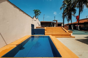 A swimming pool with steps leading up to a covered area, surrounded by a building with a tiled roof and palm trees. - Hotel Aguas do Rio Grande (Orindiúva)