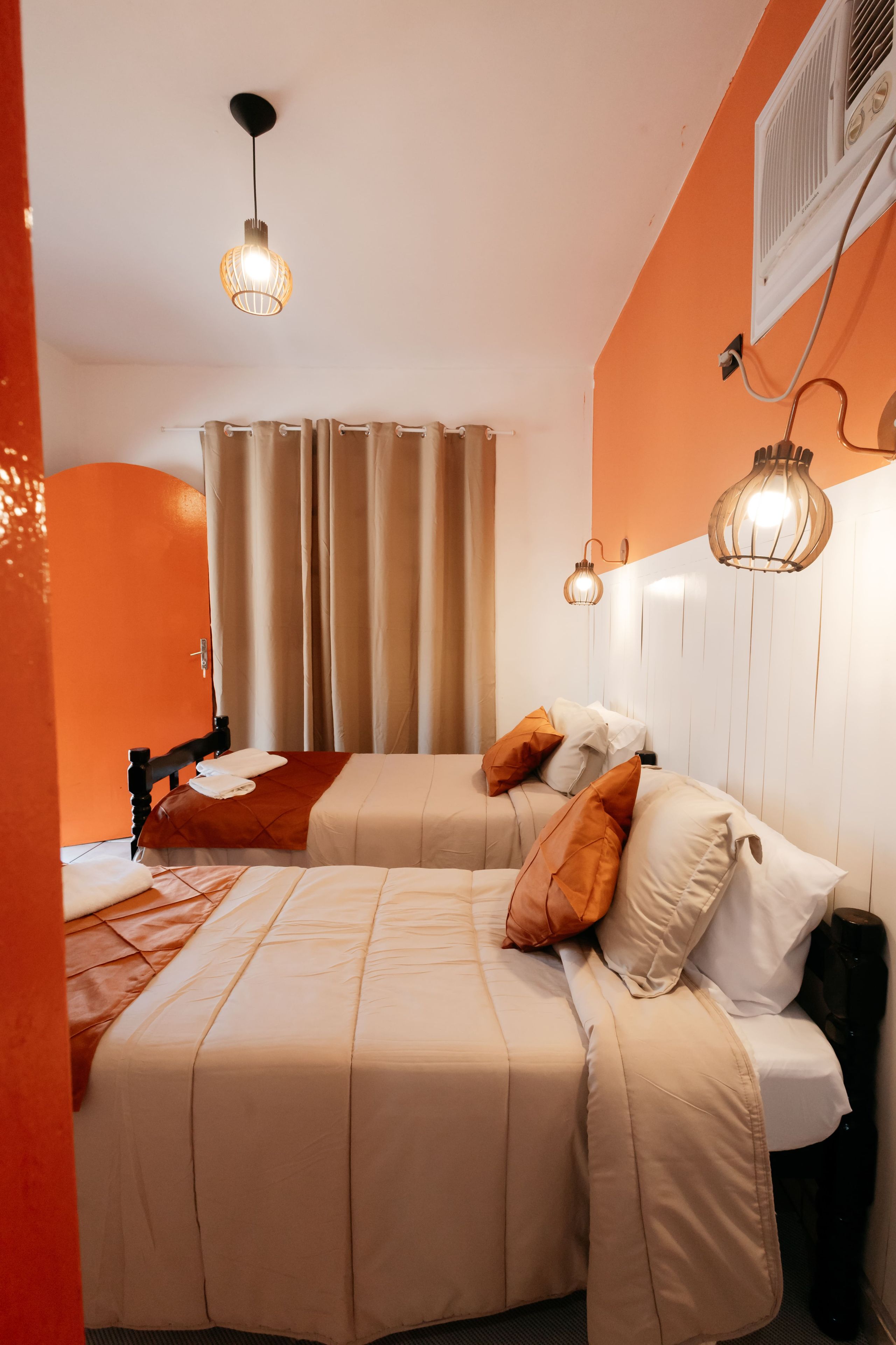 A hotel room with two beds, a window with curtains, a ceiling light, and an air conditioning unit.